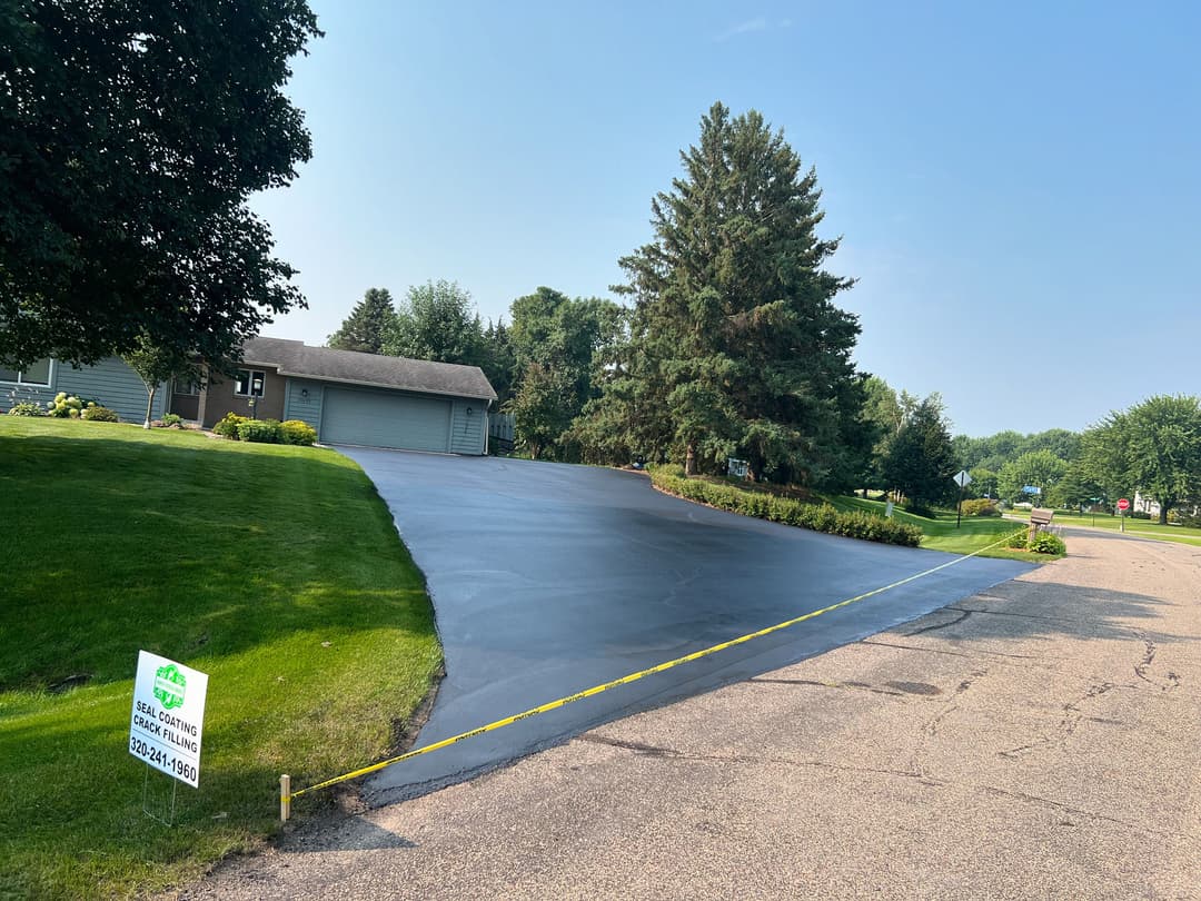 Freshly seal-coated driveway next to a home, surrounded by lush green grass and trees.