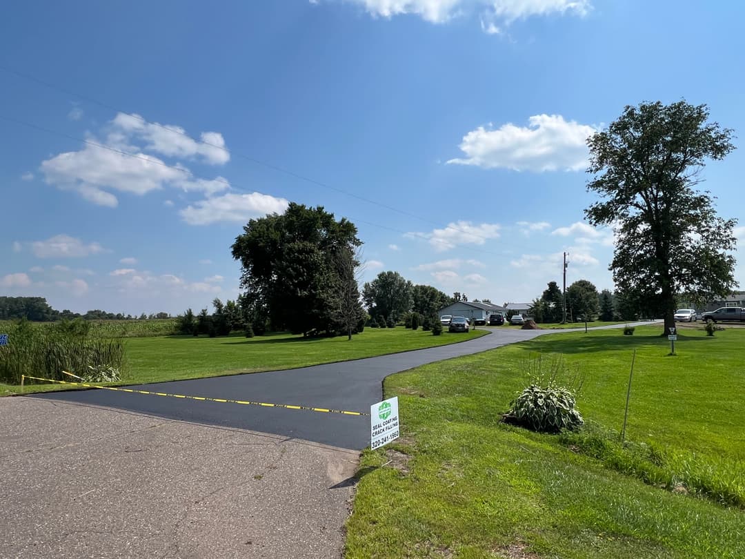 Paved road leading to a house, surrounded by green grass and trees under a clear blue sky.