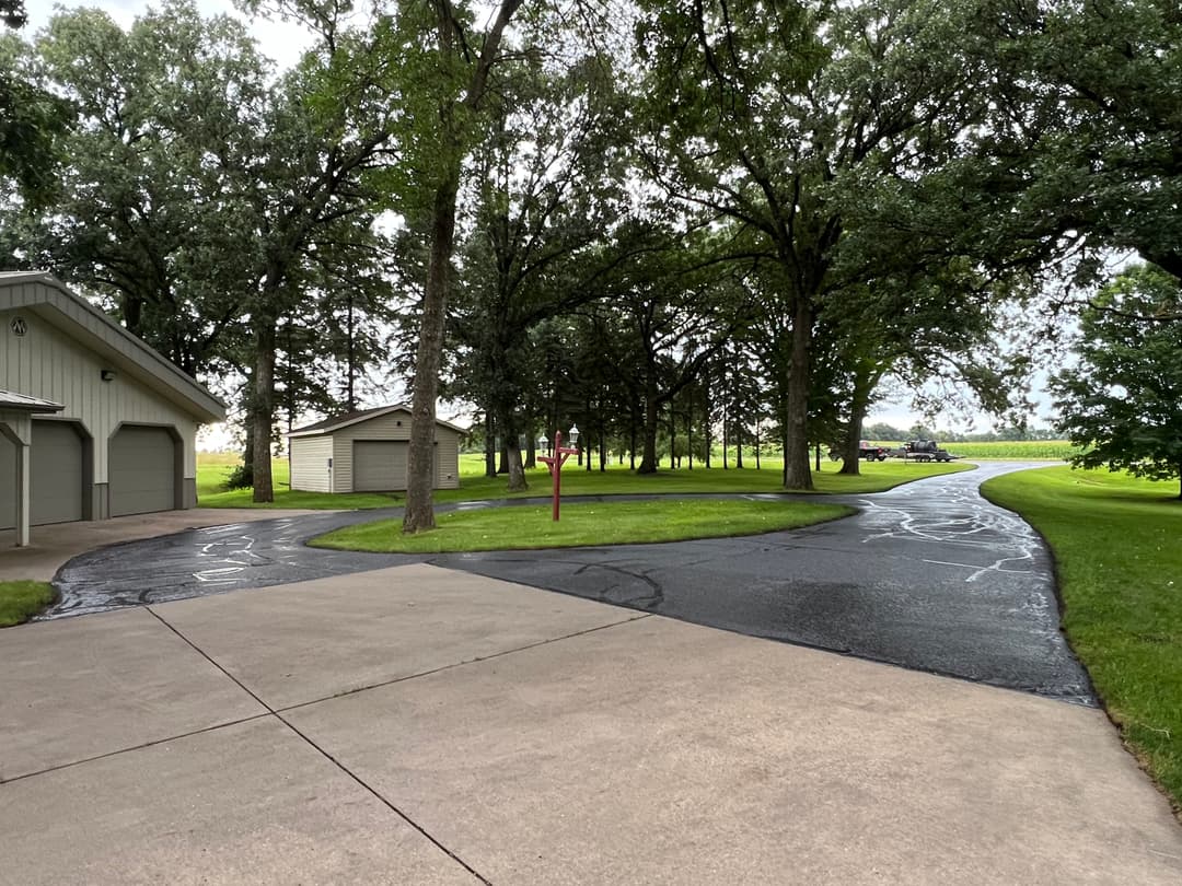 Quiet driveway winding through trees with buildings on the left and open grass on the right.