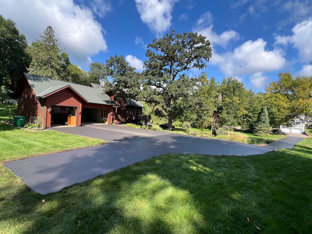 Rustic red house with green lawn and driveway under a blue sky with clouds.