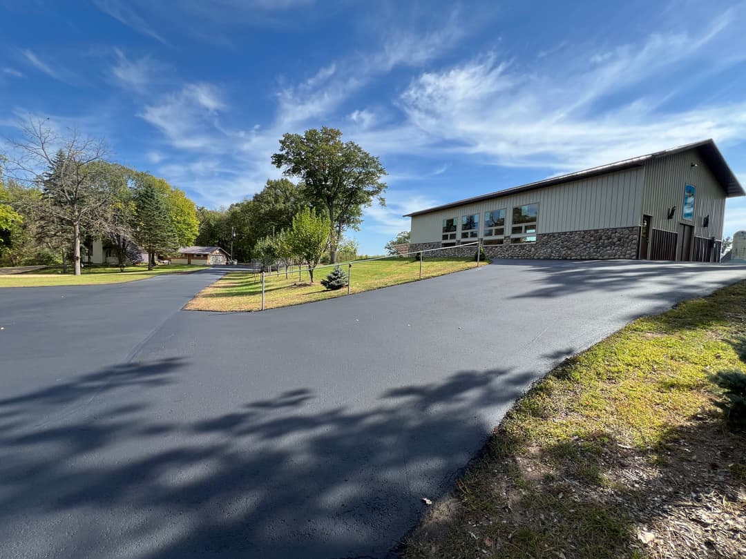 Paved driveway leading to a modern building surrounded by greenery and blue sky.