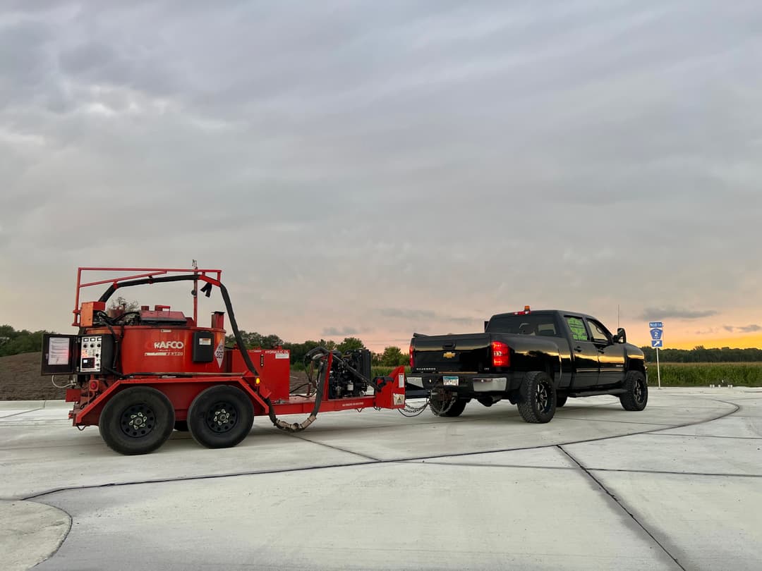 Truck towing a red asphalt repair machine on a construction site at sunset.