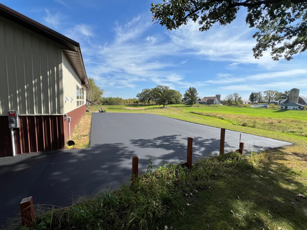 Freshly paved asphalt driveway beside a rural building and green fields under a blue sky.