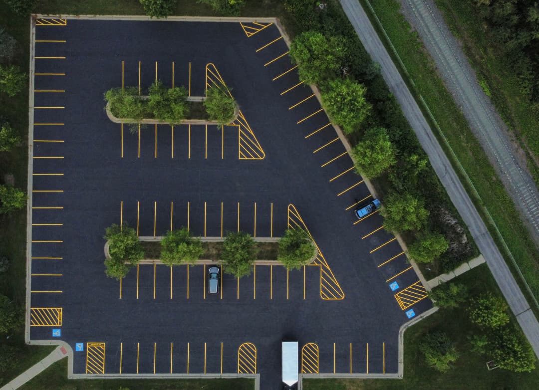 Aerial view of an empty parking lot with yellow lines and trees, near a road.