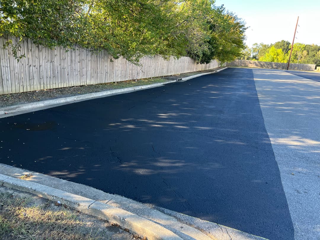 Freshly paved asphalt road beside a wooden fence and shaded trees on a sunny day.