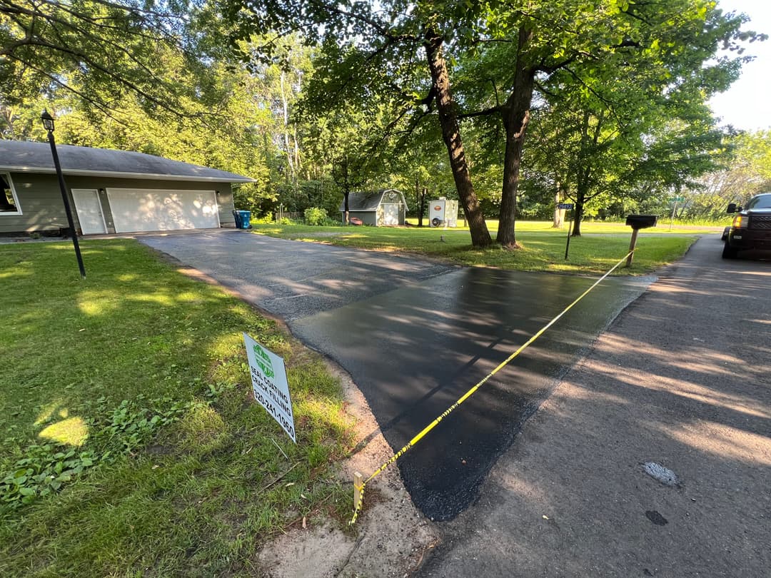 Freshly sealed driveway with measurement tape, green grass, and trees in a residential area.